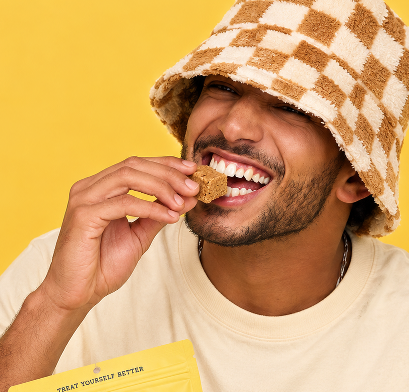 A man wearing a brown and cream checkered fuzzy bucket hat and a cream t-shirt is shown eating a brown dog treat against a solid yellow background. A yellow bag with the text 'TREAT YOURSELF BETTER' is visible in the lower left corner.