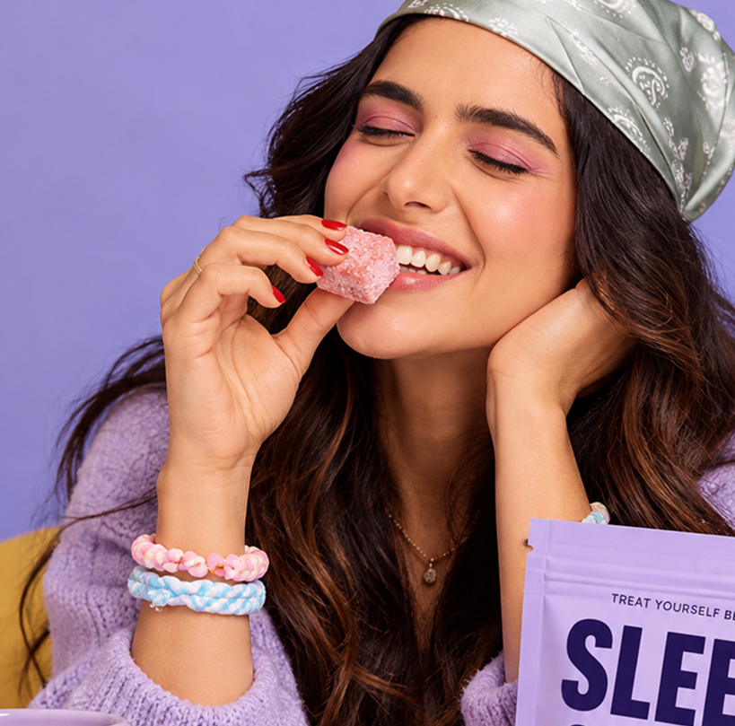 A woman wearing a light green patterned headscarf and a lavender knit sweater bites into a pink sugar-coated candy. She is wearing pastel braided hair ties on her wrist and holding a purple package labeled 'SLEEP' against a solid lavender background.